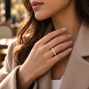 A close-up lifestyle shot of a woman wearing a delicate silver band ring set with small clear stones on her ring finger. She is wearing a beige coat, and the background is a softly blurred outdoor cafe setting.