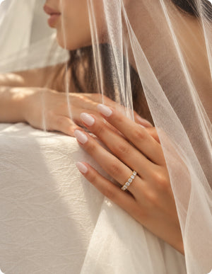 A close-up of a woman's hand with pale pink manicured nails wearing a delicate silver band ring set with small clear stones, partially veiled by sheer white tulle.