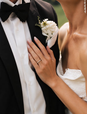 A close-up wedding scene showing a bride in a white strapless dress resting her hand on her groom's chest. The groom wears a black tuxedo jacket, white dress shirt, black bow tie, and a white floral boutonniere.