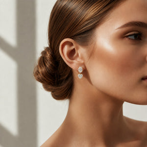 A close-up of a woman wearing a pair of small heart-shaped stud earrings with sparkling clear stones in a silver-toned setting. The image highlights her ear, jawline, and styled hair against a softly lit neutral background.