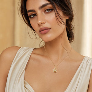 A close-up of a woman wearing a delicate gold necklace with a square pendant featuring a central clear stone, styled with a cream-colored draped top against a soft beige background.