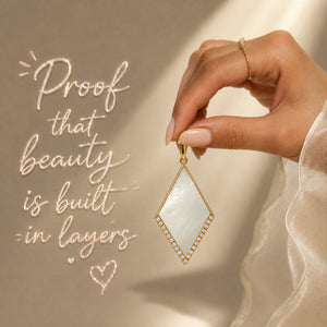 A hand wearing a delicate gold ring holds a gold diamond-shaped pendant with a white mother-of-pearl center and small clear stones along the border. The background is a warm beige with elegant cursive text that reads 'Proof that beauty is built in layers'.
