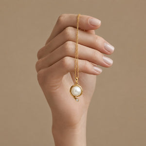 A hand with manicured nails holds a delicate gold chain necklace featuring a circular gold pendant with a white pearl and a tiny clear accent stone, set against a solid beige background.