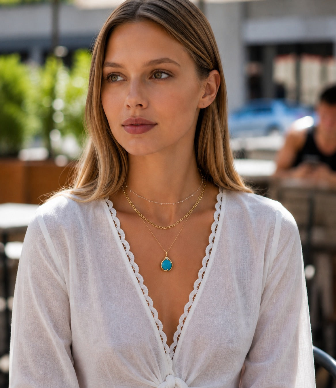 A woman wearing a white lace-trimmed long-sleeve top and layered gold necklaces, including one with a turquoise teardrop pendant, in an outdoor patio setting.