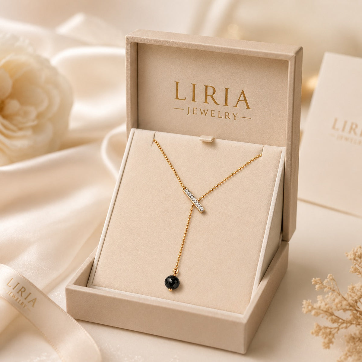 A gold Y-shaped necklace with a small clear stone and a single black bead pendant, presented in an open beige LIRIA JEWELRY box on a soft cream fabric background.