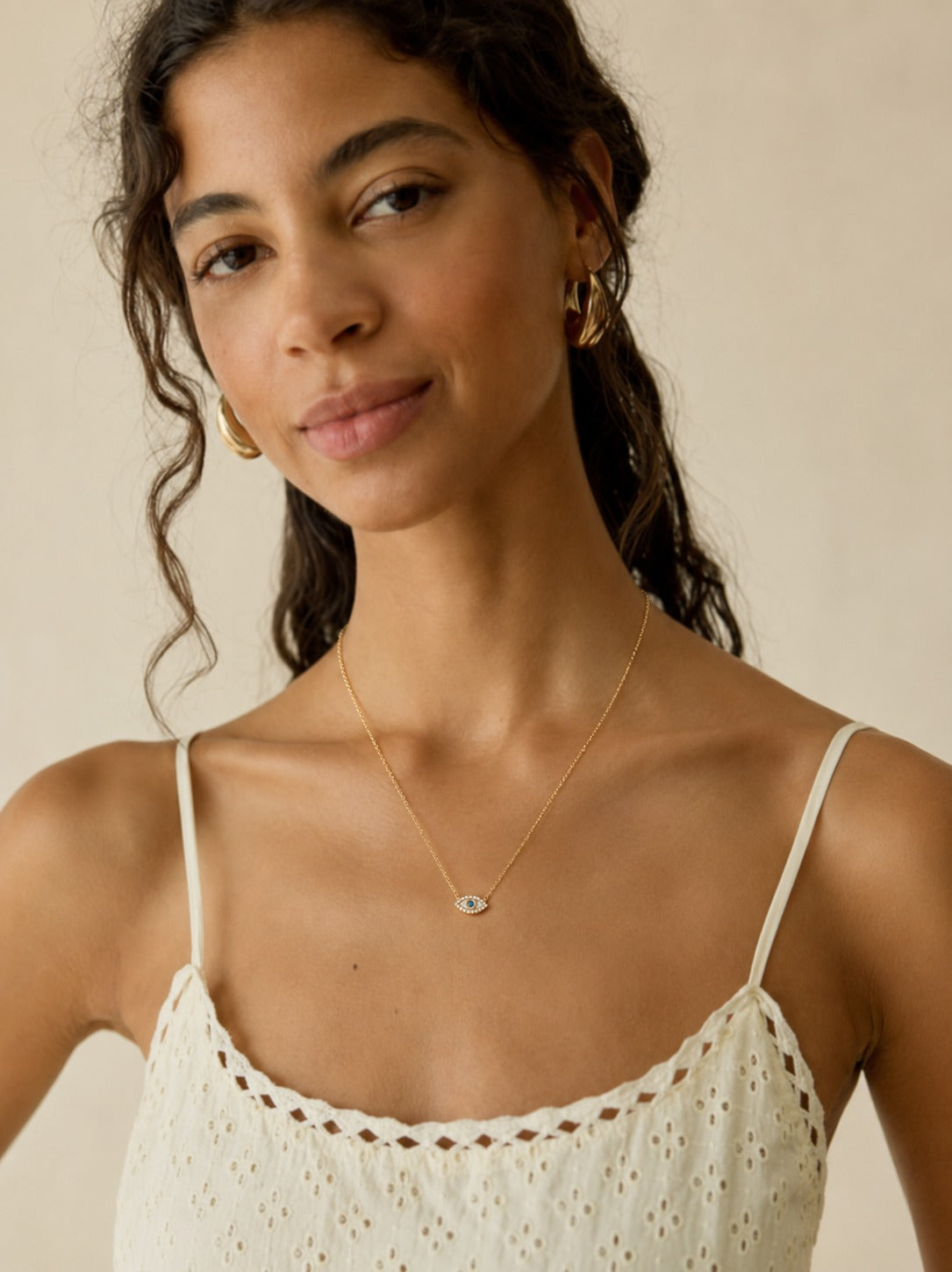 A woman wearing a delicate gold necklace with a small evil eye pendant, styled with gold hoop earrings and a white eyelet camisole top against a warm beige background.