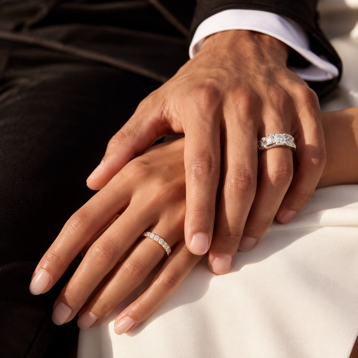 Two hands resting together, each wearing a silver band ring with clear gemstones. One hand is wearing a larger three-stone diamond engagement ring, and the other wears a smaller diamond eternity band.
