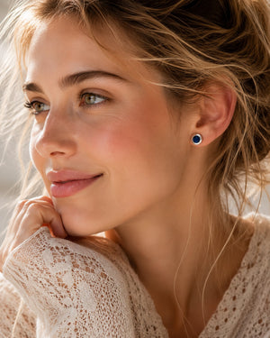 A close-up lifestyle shot of a woman with blonde hair wearing a small round stud earring with a dark blue center and a gold outer ring, paired with a cream lace top.