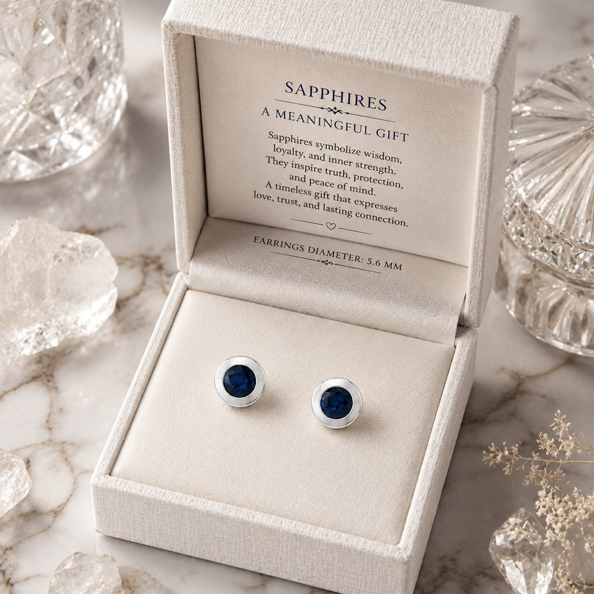 A pair of round sapphire stud earrings with silver settings displayed in an open cream jewelry box. The box lid is open, showing printed text about sapphires, and the scene is styled with clear decorative crystals on a marble surface.