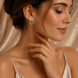 A close-up of a woman wearing a small 18k white gold hoop earring, with her hand near her neck, set against a shimmering champagne-colored fabric backdrop.
