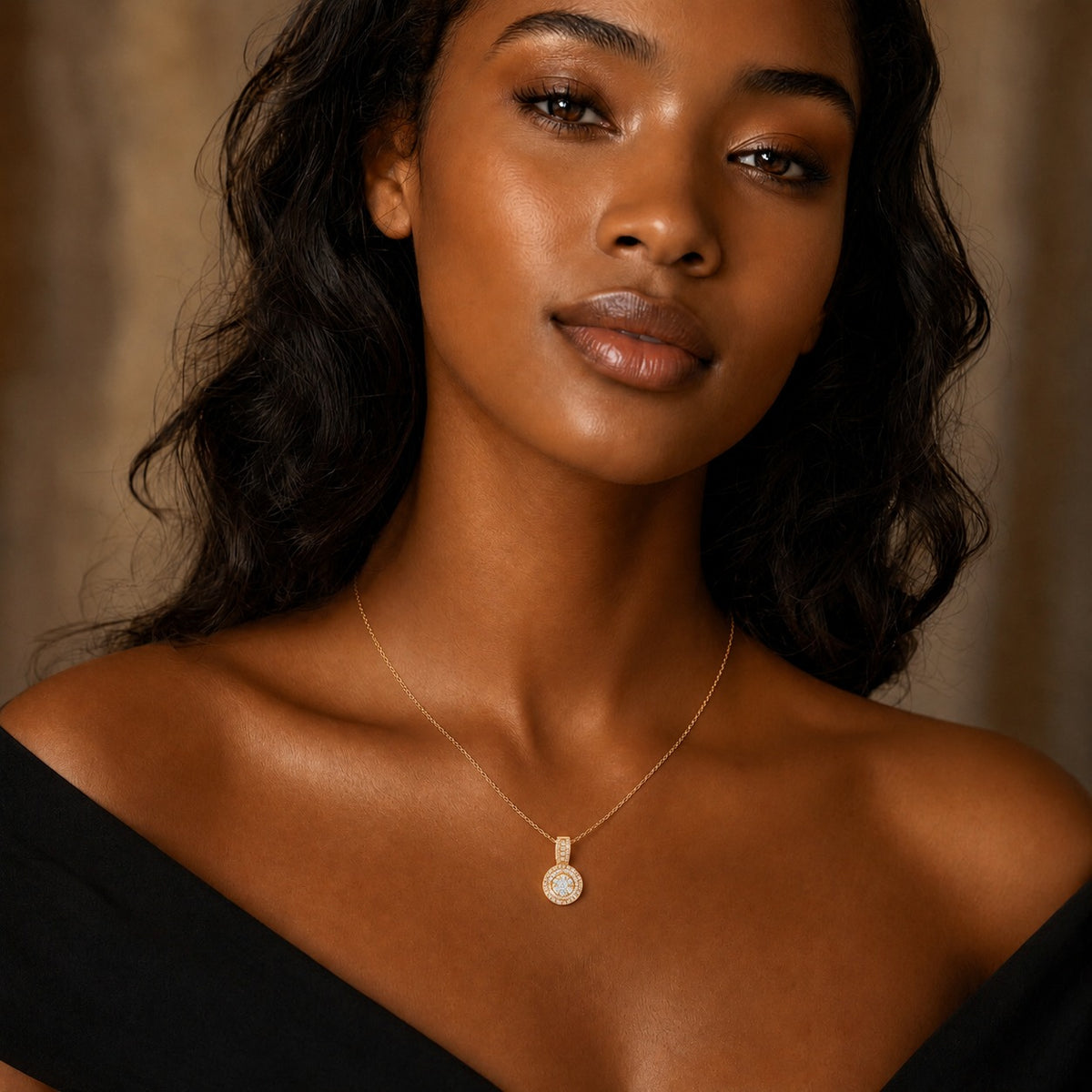 A close-up portrait of a woman wearing a delicate gold chain necklace with a small oval pendant, styled with a black off-the-shoulder top against a warm, softly blurred background.