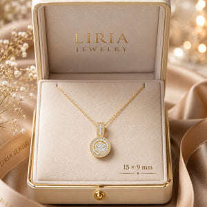 A gold pendant necklace displayed inside an open LIRIA JEWELRY box. The pendant is oval-shaped with a halo of clear stones and is shown on a cream velvet insert with size markings reading 15 x 9 mm.