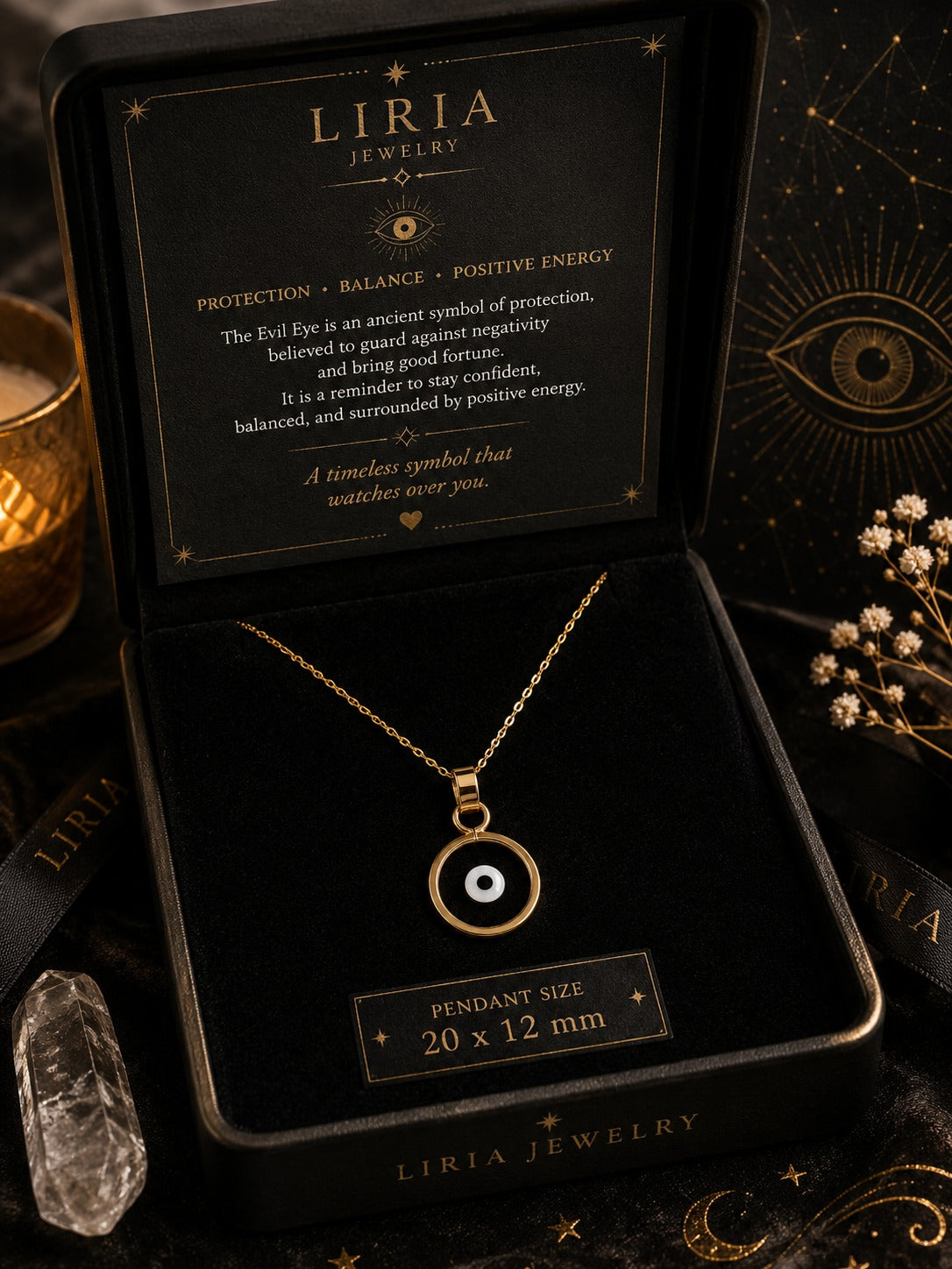 A gold-toned necklace with a circular evil eye pendant is displayed inside an open black LIRIA JEWELRY gift box. The box lid shows a black card with white text about the pendant's meaning, and a small label below the box reads 'PENDANT SIZE 20 x 12 mm'.
