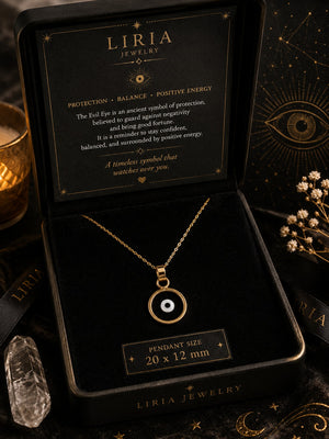 A gold-toned necklace with a circular evil eye pendant is displayed inside an open black LIRIA JEWELRY gift box. The box lid shows a black card with white text about the pendant's meaning, and a small label below the box reads 'PENDANT SIZE 20 x 12 mm'.