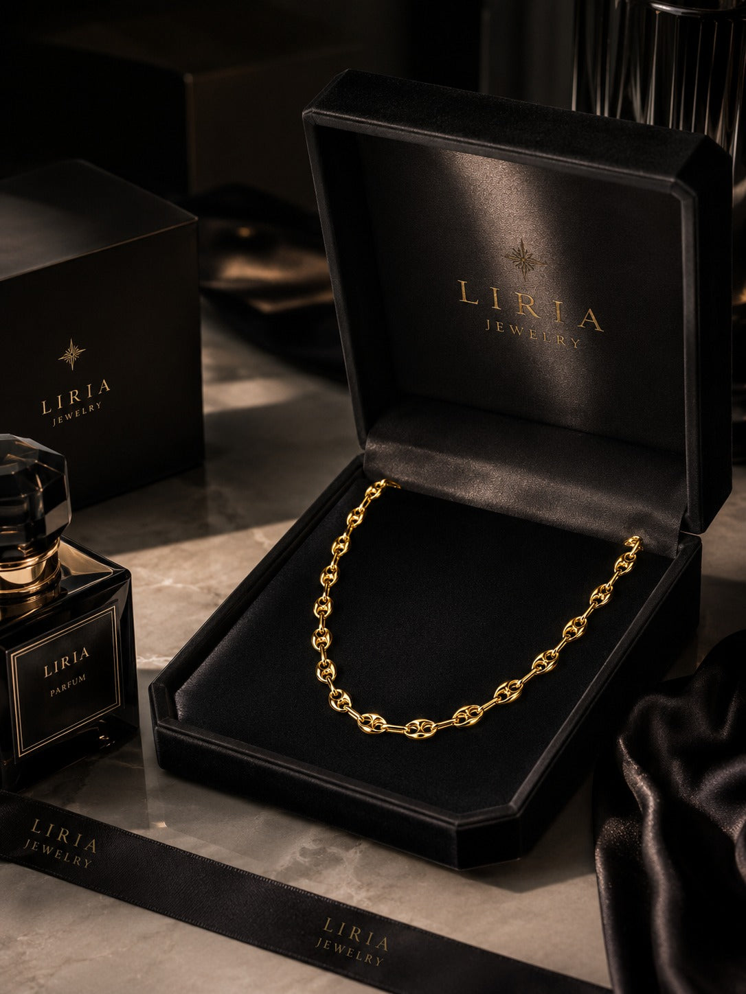 A gold chain necklace displayed inside an open black LIRIA JEWELRY box, with matching branded packaging and a perfume bottle on a dark marble surface.