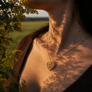 A close-up lifestyle shot of a woman outdoors wearing a delicate gold heart pendant necklace. The warm golden hour sunlight casts leaf shadows across her skin, with a blurred green field and yellow flowers in the background.