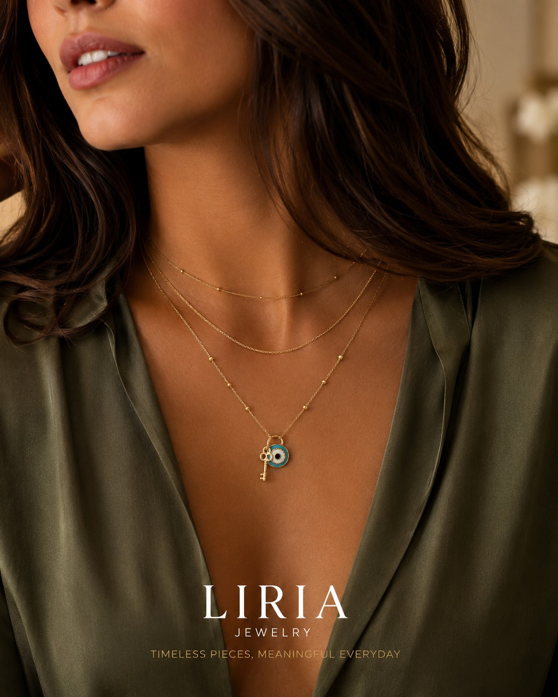 A close-up lifestyle shot of a woman wearing a layered gold necklace with a key and evil eye pendant, styled with a beige satin shirt.