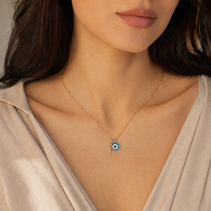 A close-up of a woman wearing a delicate gold chain necklace with a small evil eye pendant. The necklace features tiny gold beads along the chain and a central blue and white evil eye charm with a small gold key detail.