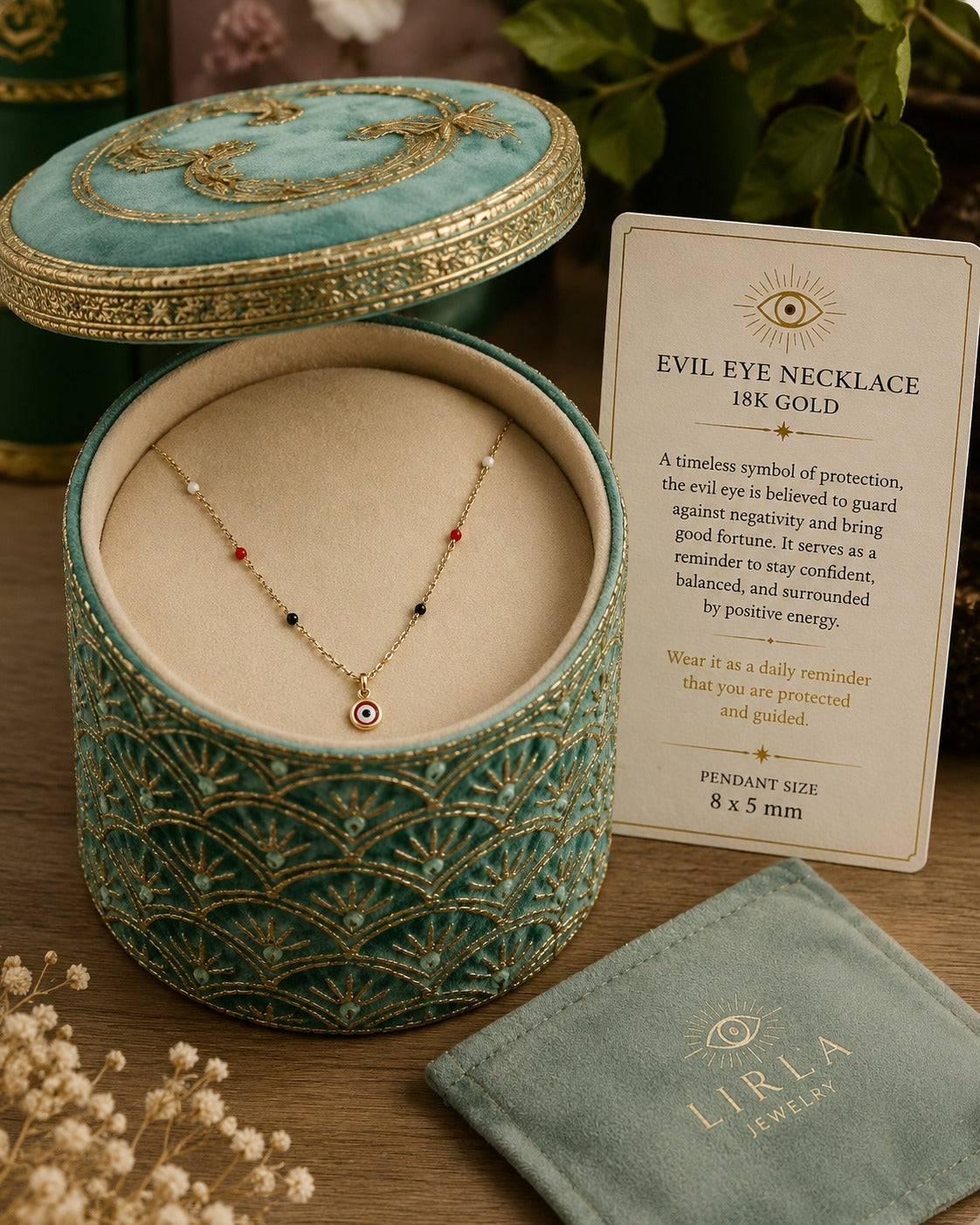 A gold necklace with a small evil eye pendant is displayed inside an open teal jewelry box with gold detailing, alongside a product card and a branded pouch on a wooden surface.
