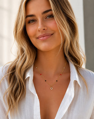 A close-up of a woman with blonde hair wearing a delicate gold necklace with small red and black beads and a circular pendant, paired with a white collared shirt.
