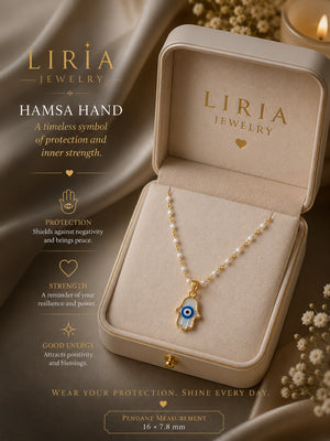 A Liria Jewelry Hamsa hand pendant necklace displayed in an open beige jewelry box, with decorative draped fabric, dried flowers, and a lit candle in the background. Text on the left describes the protection and symbolism of the Hamsa hand pendant.