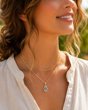 A close-up lifestyle shot of a woman wearing a double-layered gold necklace with a blue and white evil eye hamsa pendant, set against a soft, blurred outdoor background.