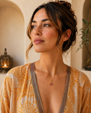 A woman wearing a delicate gold necklace with a small red and white evil eye pendant, paired with a yellow and white patterned bohemian-style top, set against a warm, softly lit background with arches.