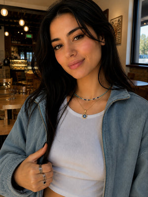 A woman with long dark hair wearing a light blue corduroy zip-up jacket over a white ribbed crop top, posing in a cafe setting.