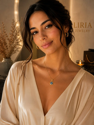 A woman wearing a delicate gold chain necklace with a blue and white evil eye pendant, styled with a cream satin top in a warm, softly lit jewelry boutique setting.