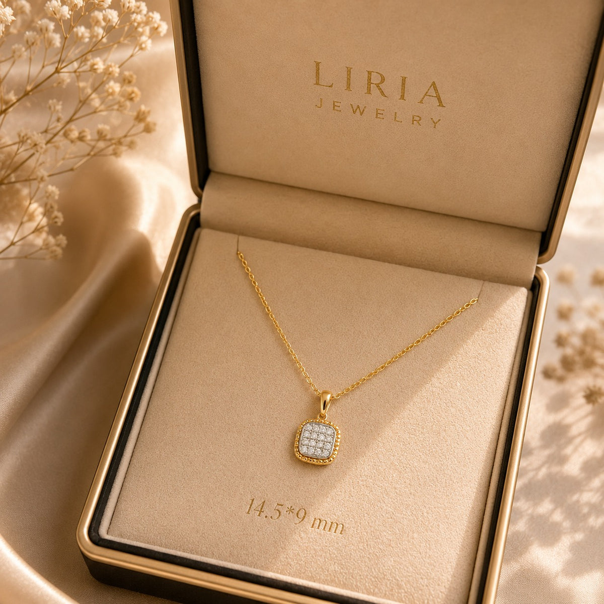 A gold pendant necklace with a square pavé-set clear stone center, displayed inside an open LIRIA JEWELRY box on a beige satin-like surface with dried flowers around it.