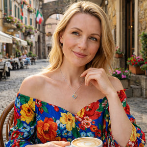 Woman in a colorful floral dress sitting outdoors in a charming town square, holding a cup of coffee.