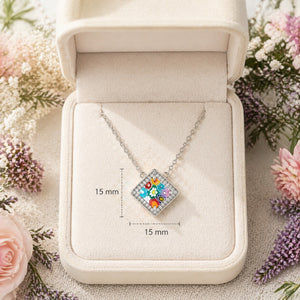 Necklace with a colorful square pendant in an open jewelry box surrounded by flowers.