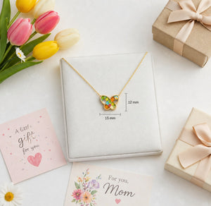 Butterfly-shaped necklace with colorful stones in a jewelry box, surrounded by flowers and cards on a white surface.