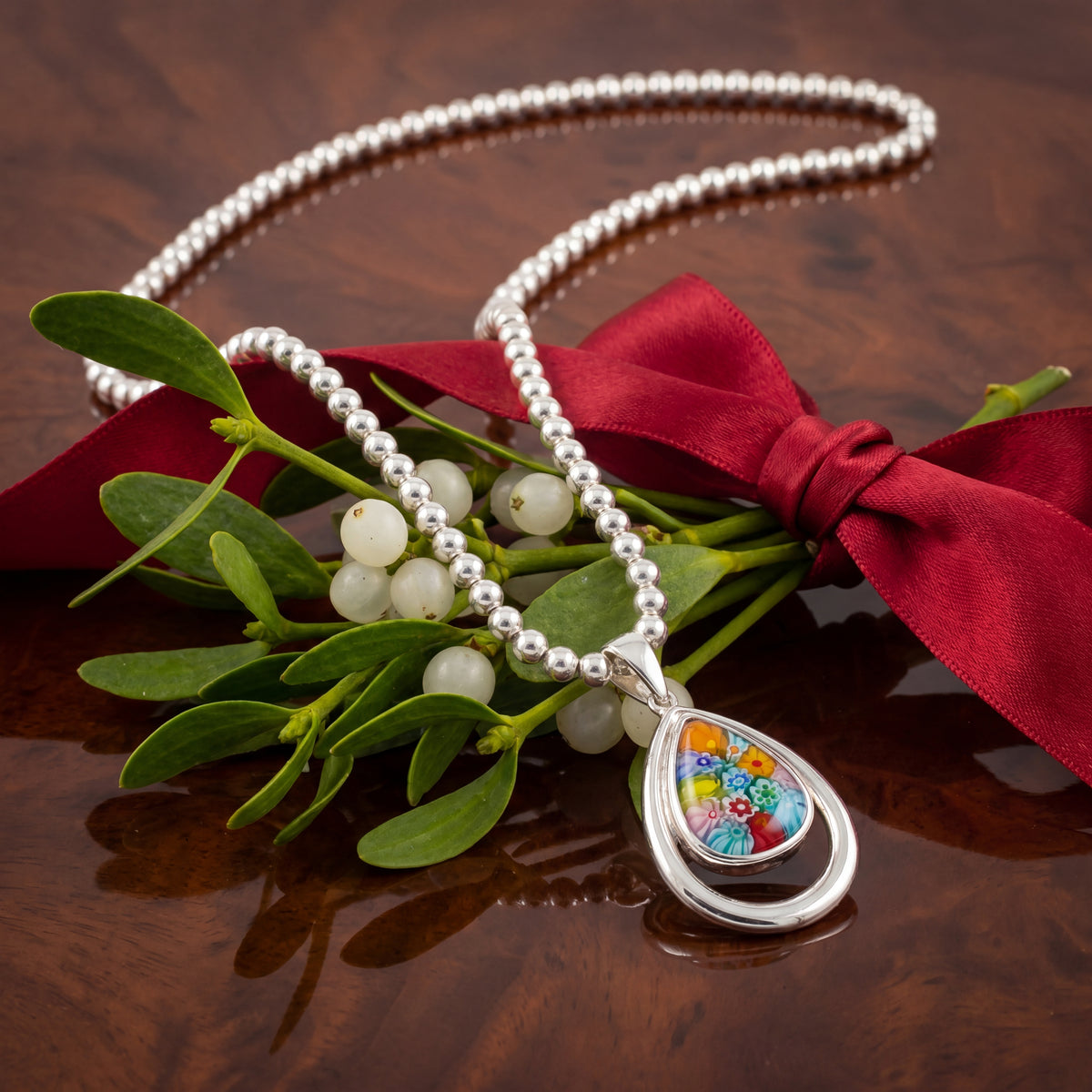Silver necklace with a colorful pendant on a wooden surface with mistletoe and a red bow.