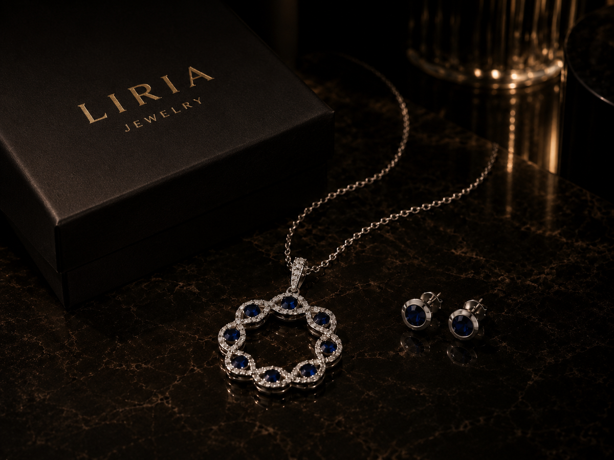 A Liria Jewelry set crafted in 14K white gold, featuring a pendant necklace and matching stud earrings accented with deep blue gemstones, elegantly displayed on a dark marble surface beside a black Liria Jewelry branded jewelry box.
