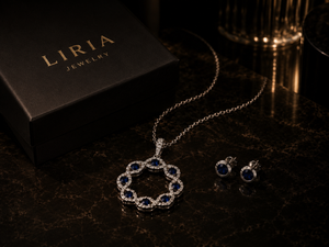 A Liria Jewelry set crafted in 14K white gold, featuring a pendant necklace and matching stud earrings accented with deep blue gemstones, elegantly displayed on a dark marble surface beside a black Liria Jewelry branded jewelry box.