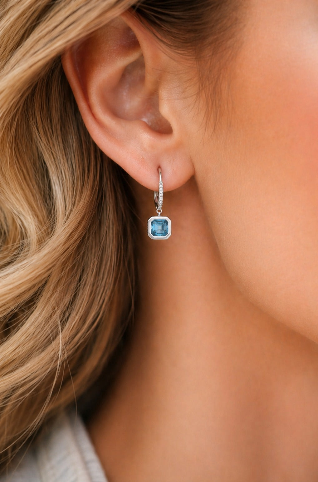 Close-up of an ear wearing a blue gemstone earring with blonde hair.