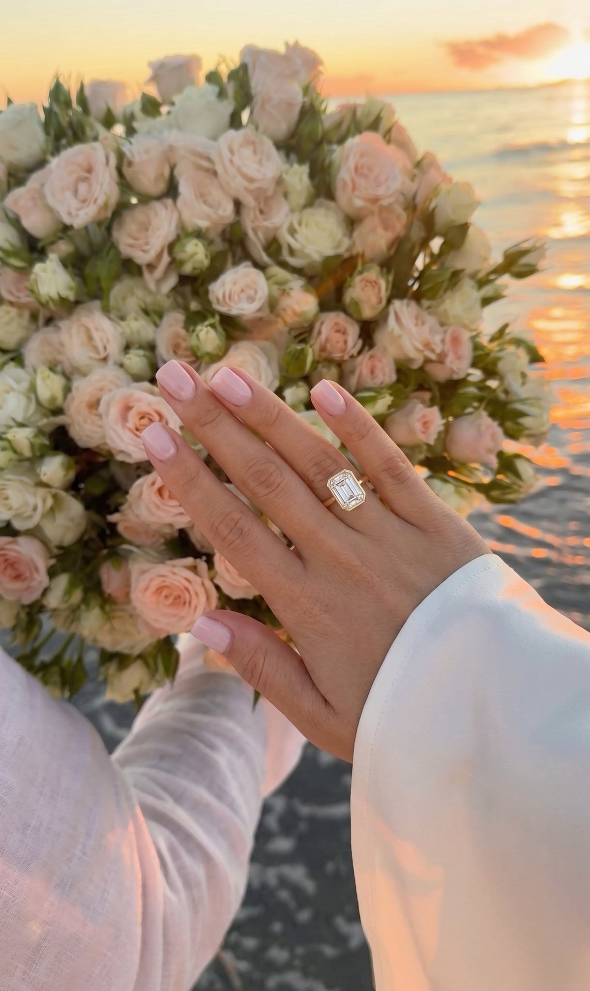 A close-up of a hand wearing a diamond engagement ring, resting near a bouquet of pale pink and white roses, with an ocean sunset in the background.