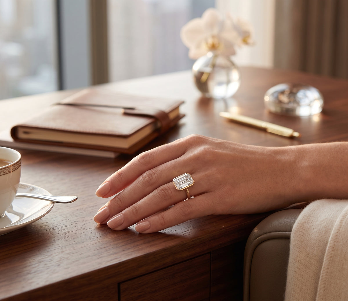 A close-up lifestyle shot of a hand resting on a wooden desk, wearing a gold ring with a large rectangular emerald-cut diamond. The background features a warm, softly blurred office or study setting with a notebook, pen, and a cup of tea.