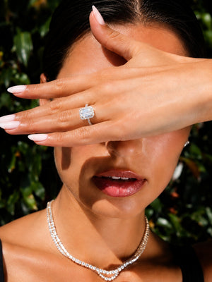 A close-up lifestyle shot of a model wearing a diamond ring on her finger, with her hand covering part of her face. She is also wearing a diamond necklace, set against a dark green foliage background.