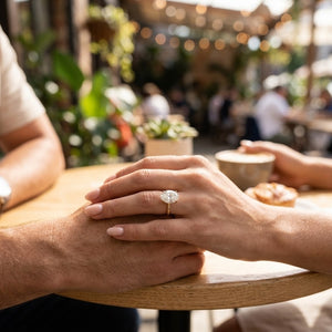 A close-up lifestyle shot of a woman's hand resting on a man's at an outdoor cafe table, wearing a gold engagement ring with a round brilliant-cut diamond.