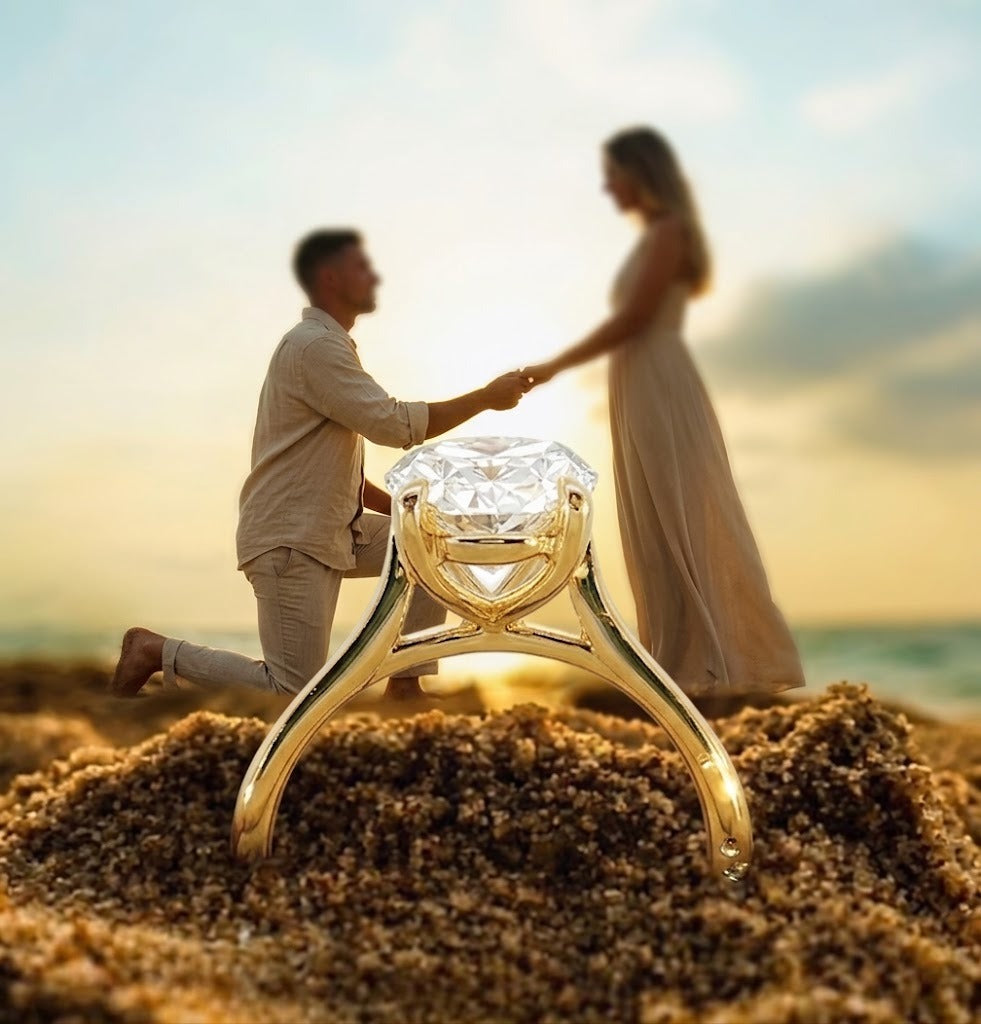 A large gold engagement ring with a clear gemstone is shown in sharp focus in the foreground, resting on sand, while a blurred couple holds hands in the background on a beach at sunset.