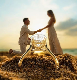 A large gold engagement ring with a clear gemstone is shown in sharp focus in the foreground, resting on sand, while a blurred couple holds hands in the background on a beach at sunset.