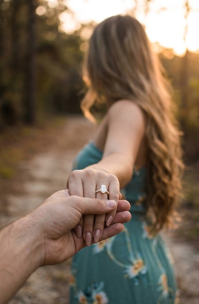A close-up of a hand holding another person's hand outdoors at sunset, with a ring featuring a clear gemstone and a small gold band worn on the finger. The person is wearing a teal floral dress, and the background shows a blurred wooded path.