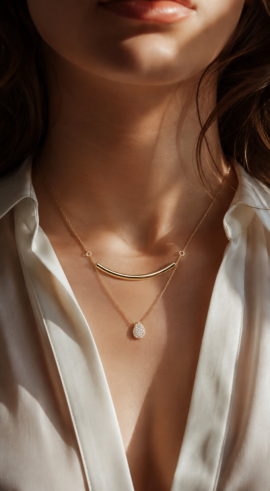 A close-up of a woman wearing a delicate gold necklace with a curved bar and a small textured oval pendant, styled with a white satin shirt under warm, dramatic sunlight.