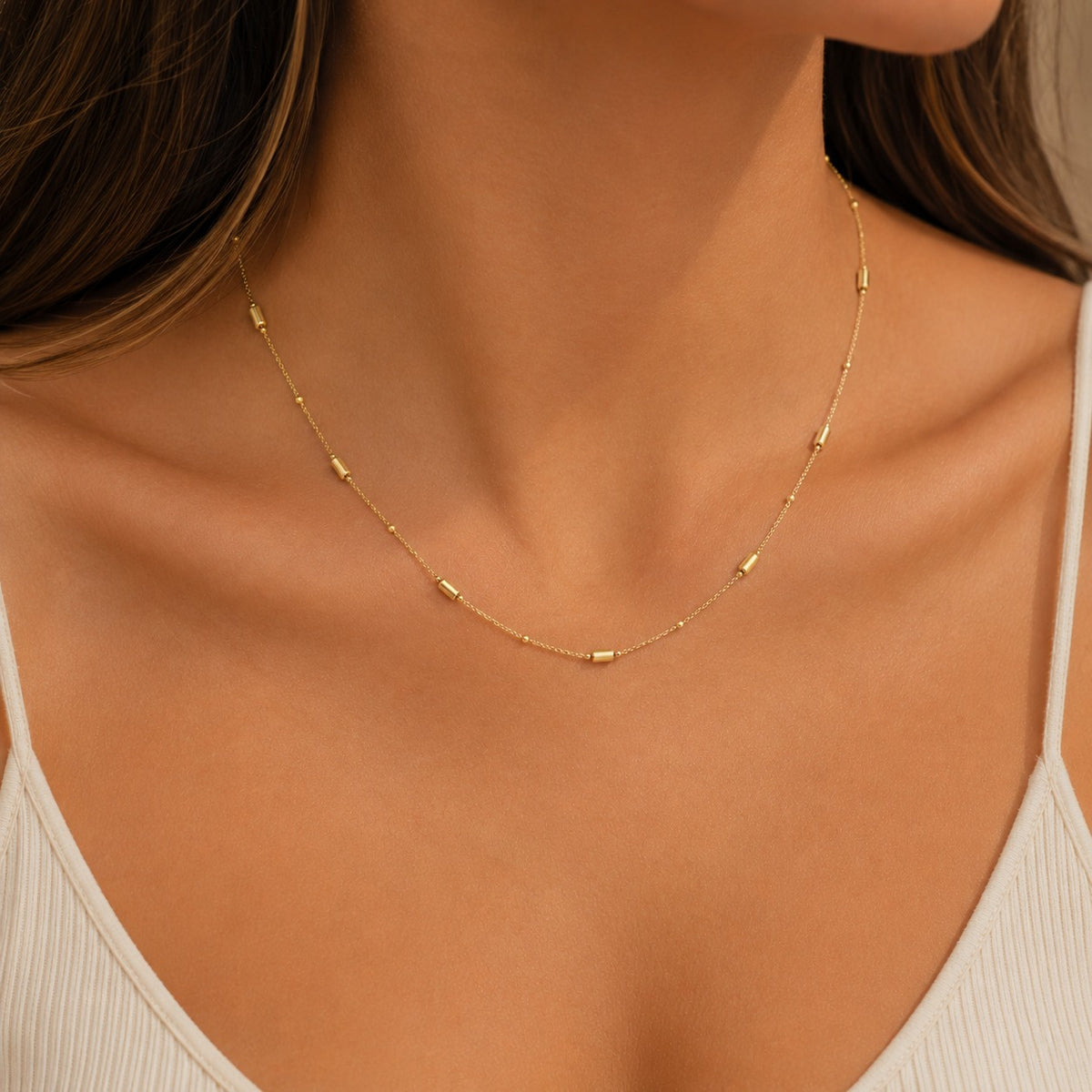 A delicate gold chain necklace with small cylindrical beads, worn on a model's neck over a white ribbed tank top.