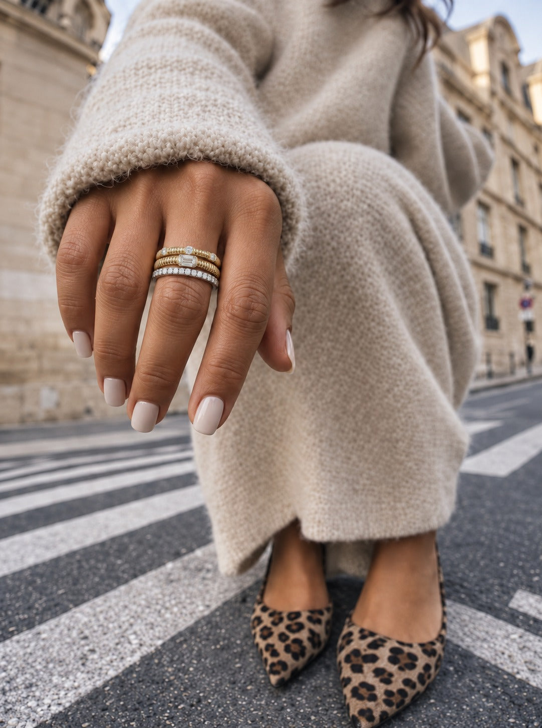 A close-up lifestyle shot of a person wearing two stacked rings on their hand. One ring is a gold band with a central clear stone, and the other is a silver band with a row of small clear stones.