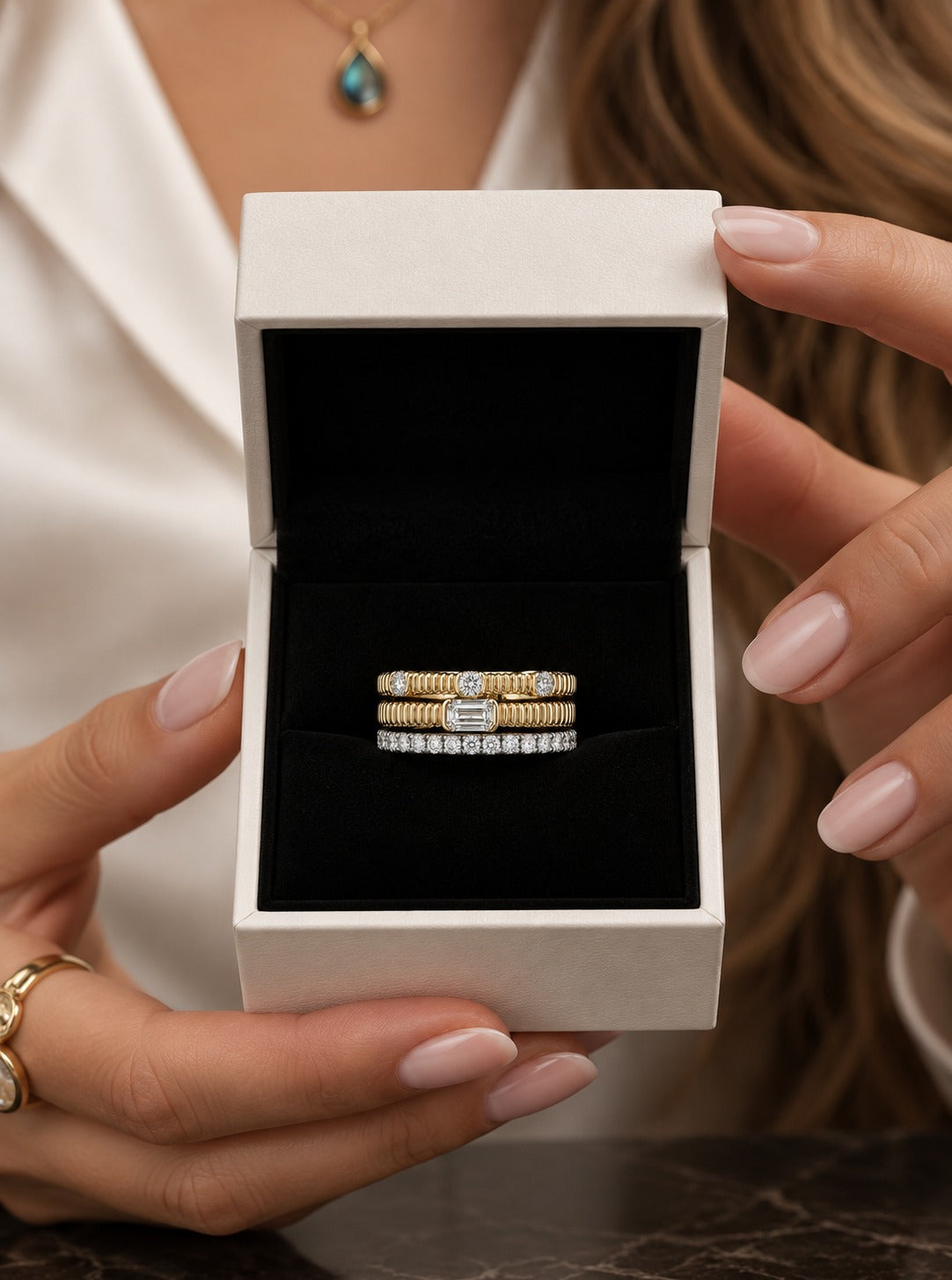 A close-up of a person holding an open white jewelry box with a black interior, displaying two slim rings. The person wears a white satin top and a teardrop pendant necklace.