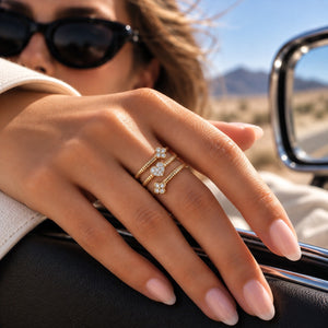 A close-up of a person's hand wearing two gold rings with small white floral clusters, photographed outdoors near a car window with sunglasses and a desert landscape in the background.