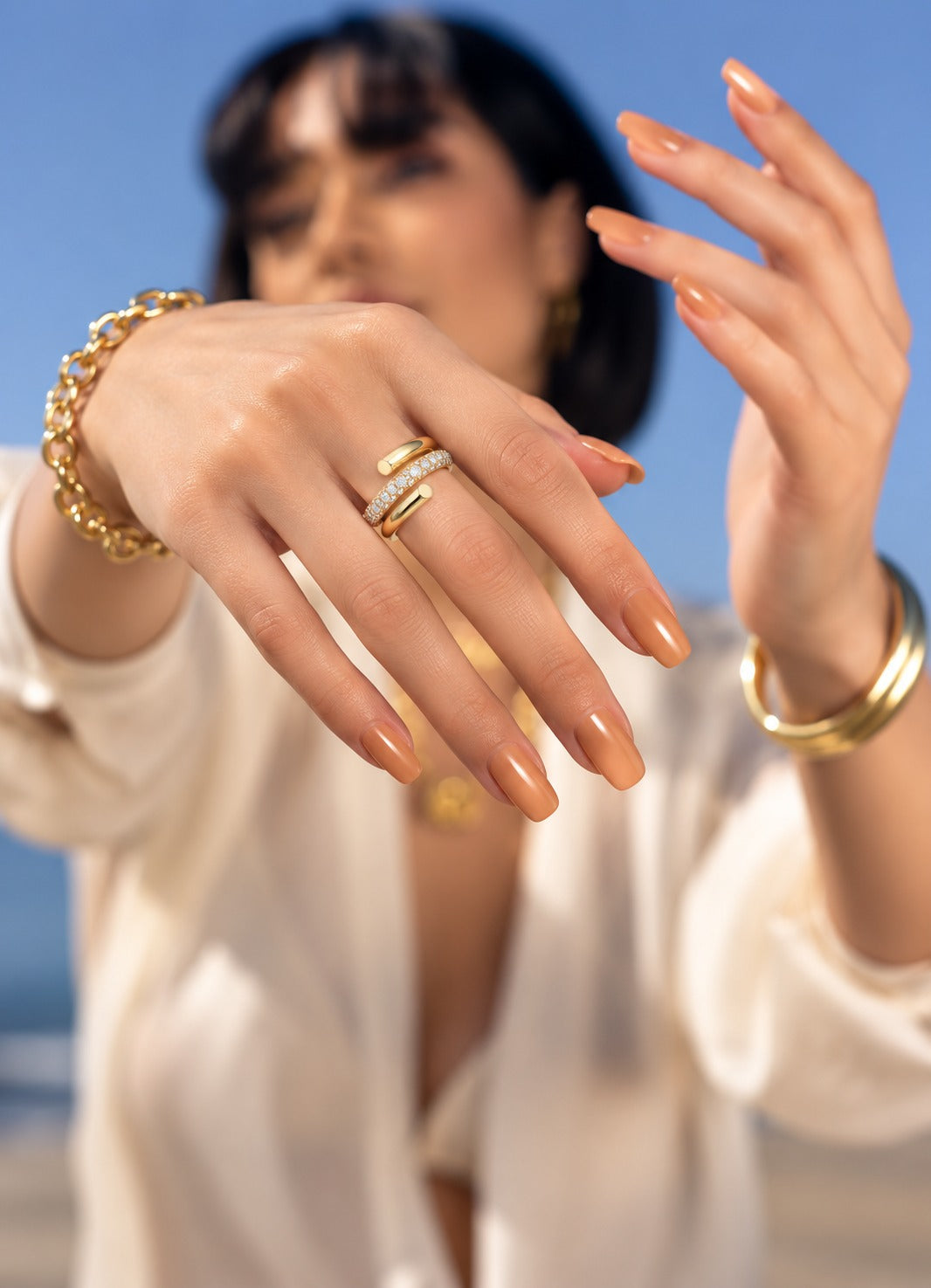 A close-up lifestyle shot of a model wearing gold rings and bracelets, with her hands raised in front of a soft-focus blue sky background.